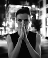 Woman in black and white with hands pressed, city lights background.