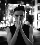 Woman in black and white with hands pressed, city lights background.
