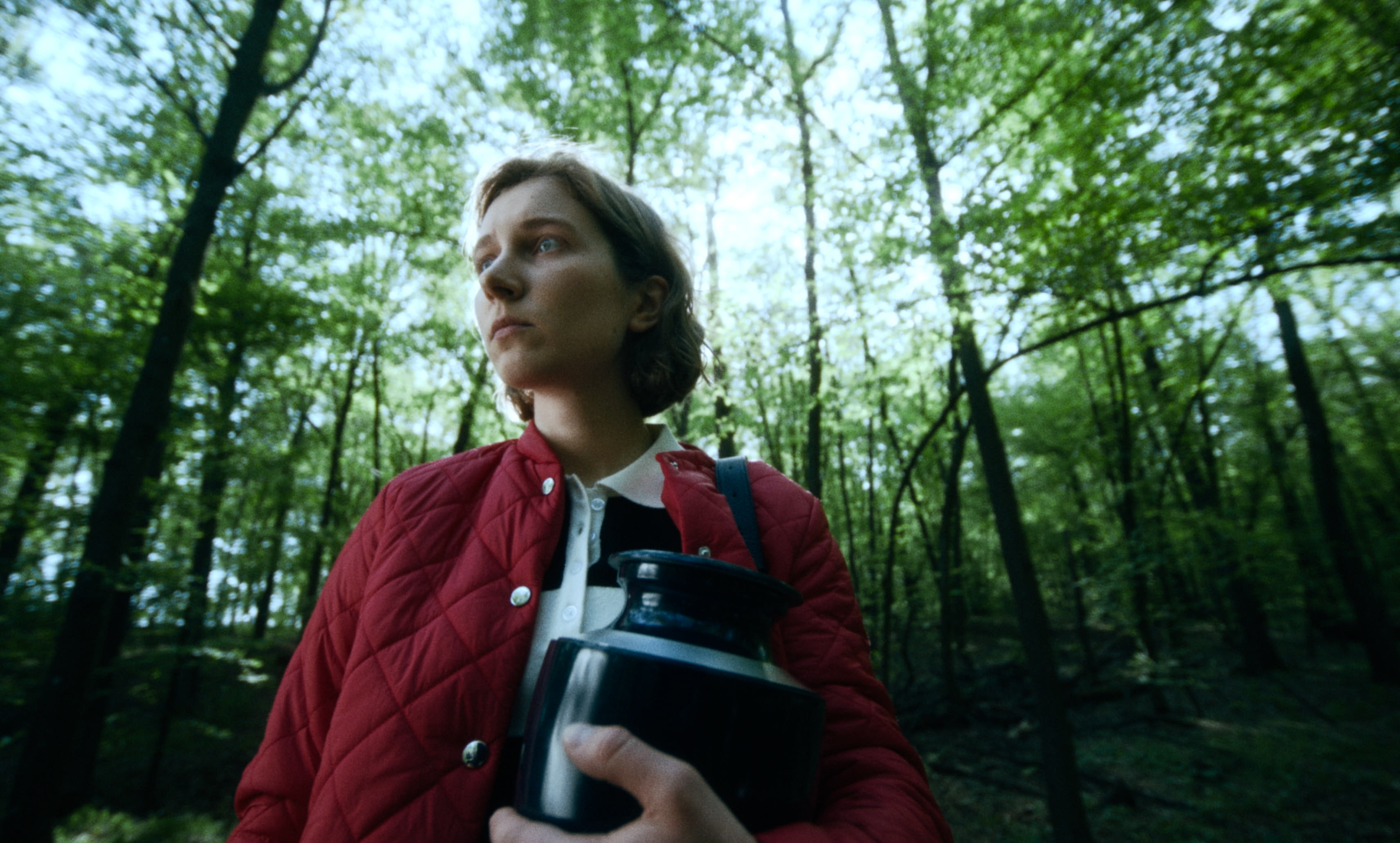 A young woman in a red jacket walks through a forest holding an urn — a still from Kira’s Dream.