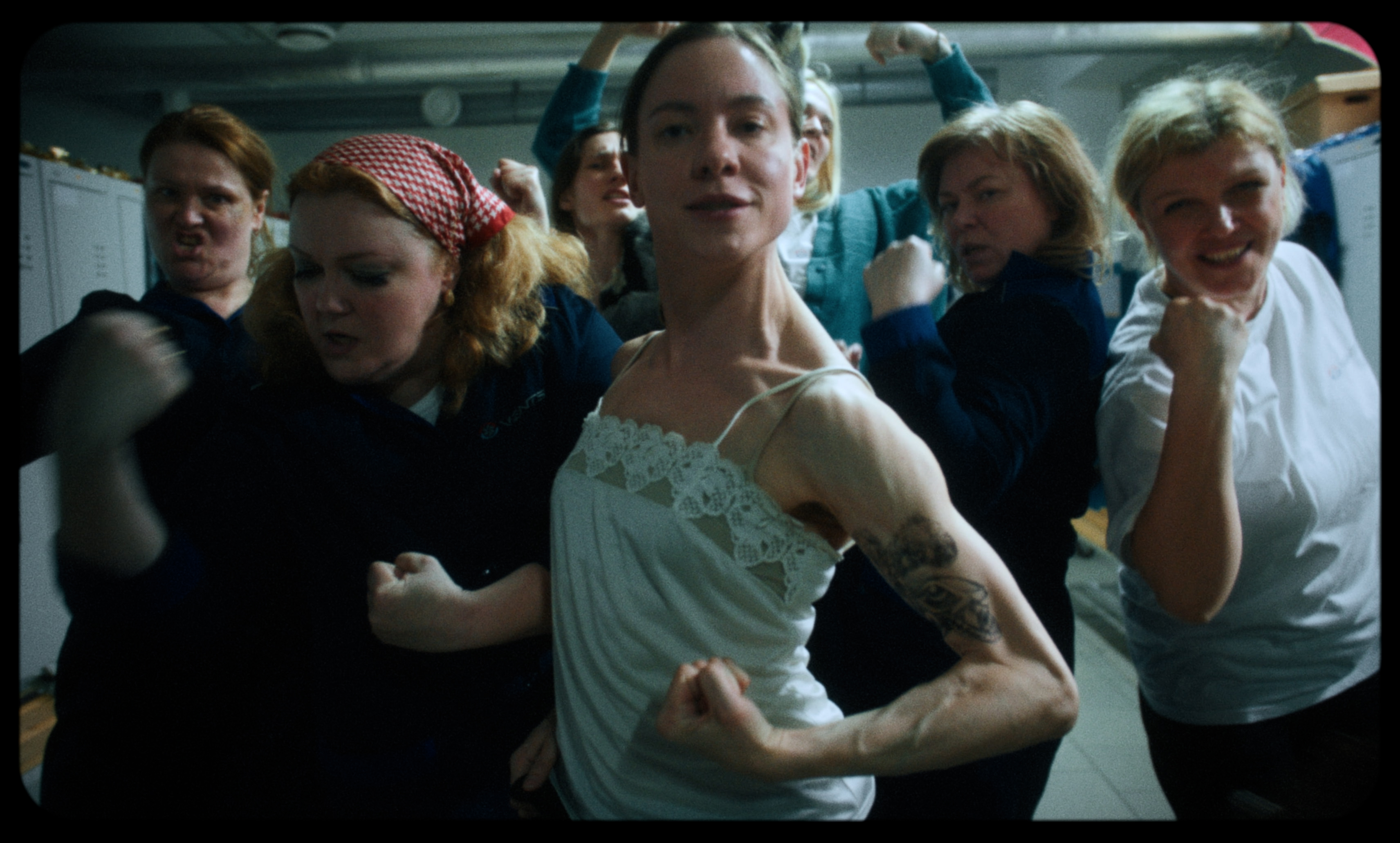 Workers pose with raised fists in a locker room, led by a young man in a slip — a still from Kira’s