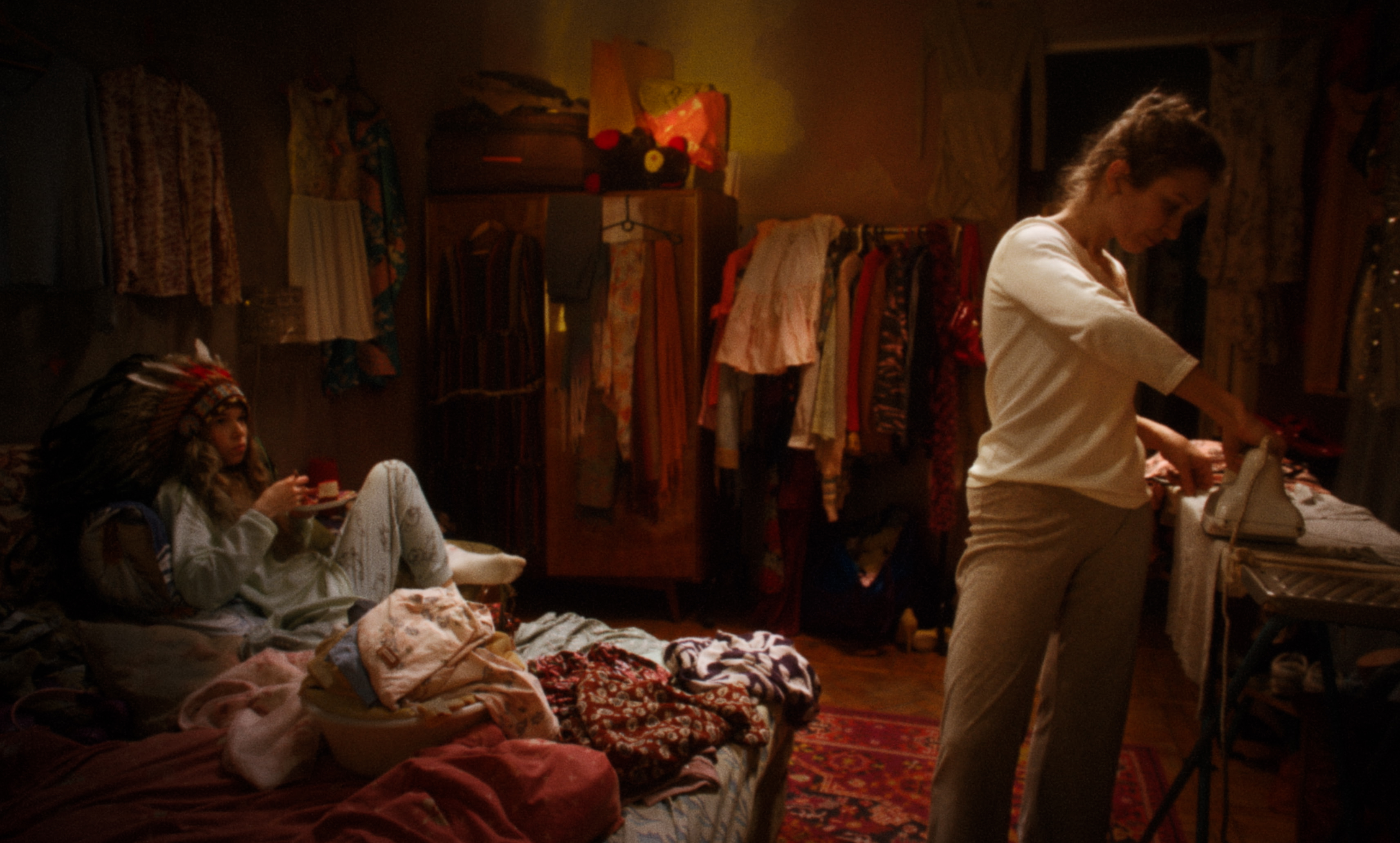 A young woman irons clothes in a cluttered bedroom while another lounges nearby — a still from Kira’