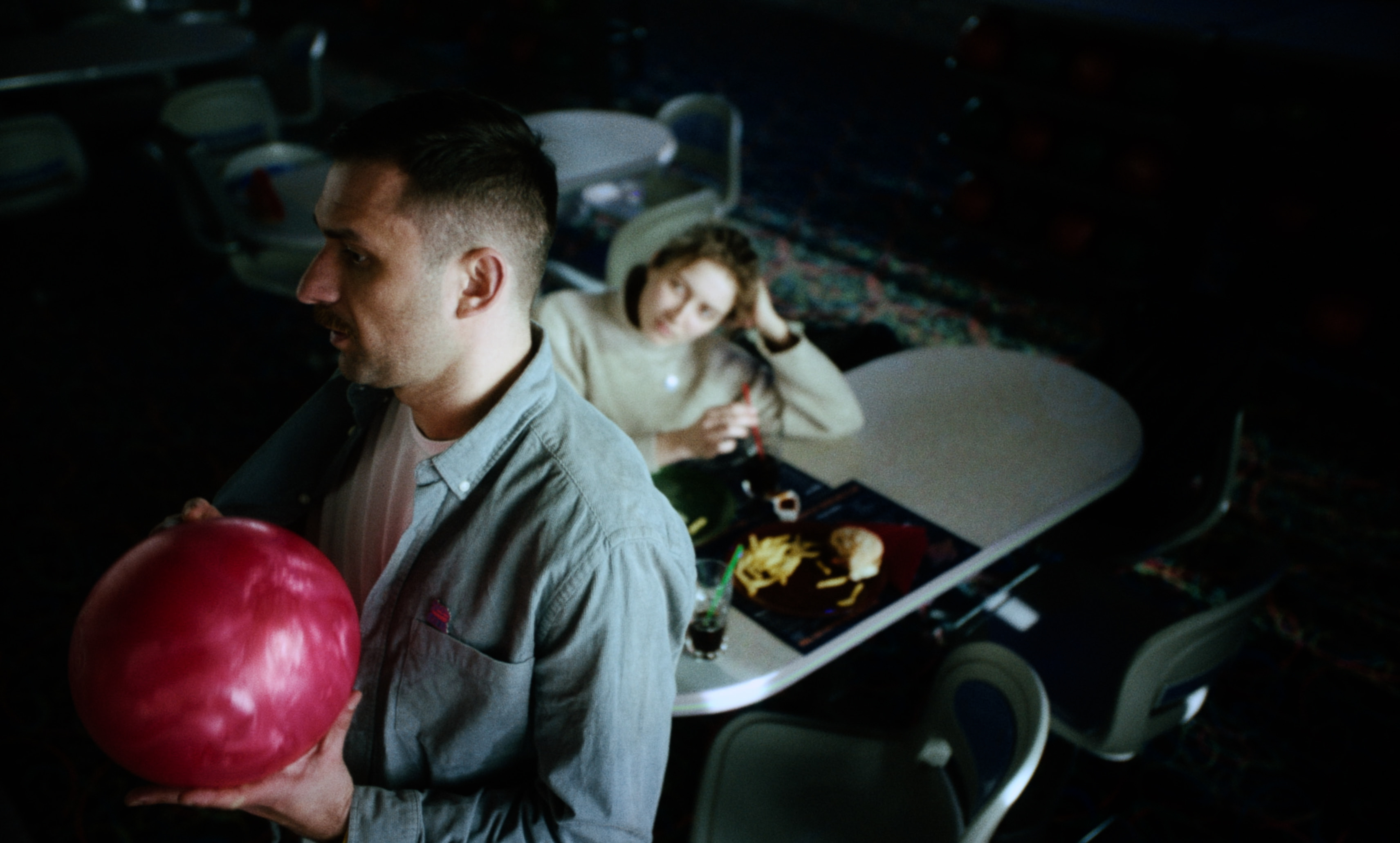 A man holds a pink bowling ball while a young woman watches from a table — a still from Kira’s Dream
