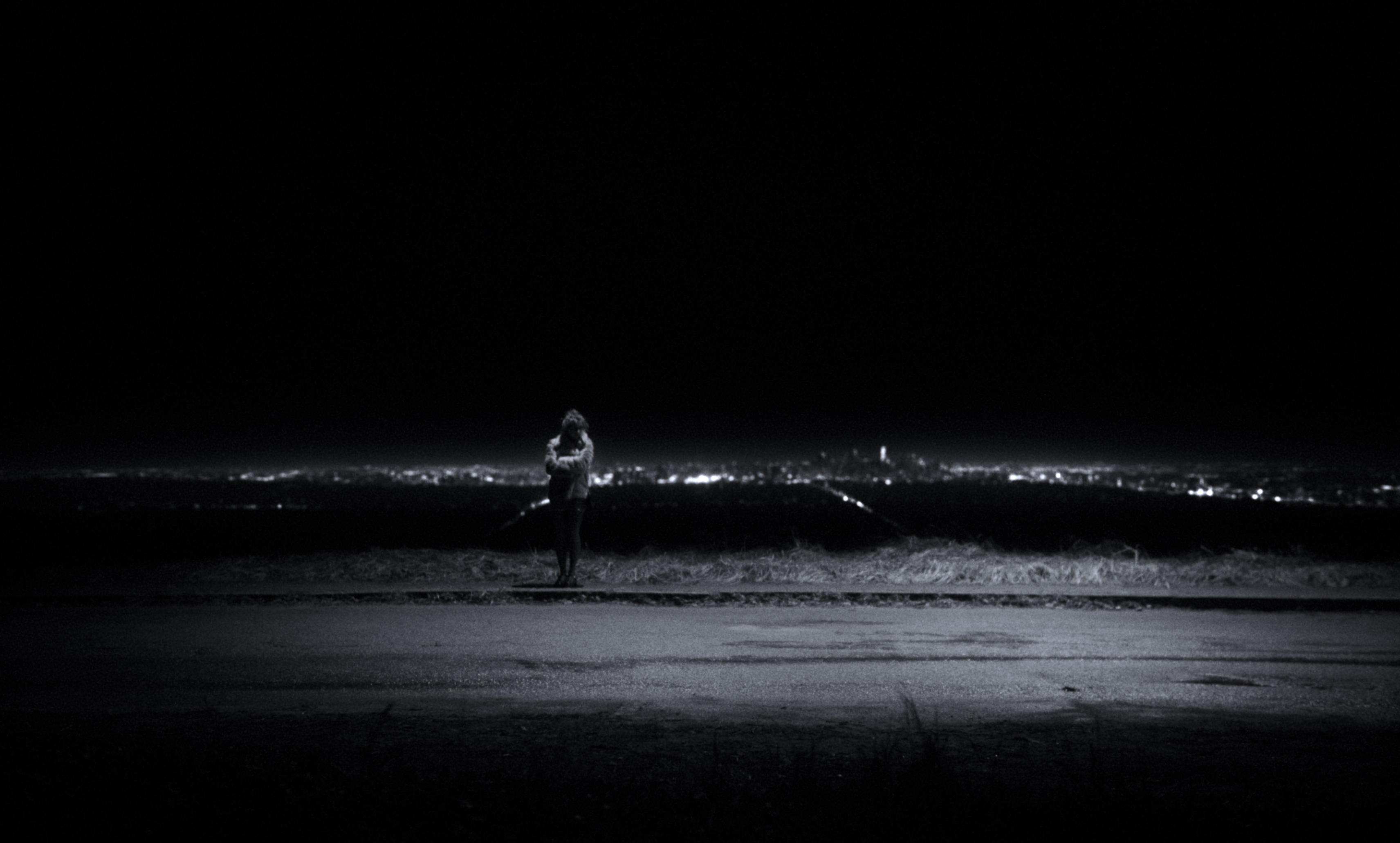 A lone figure stands at night above distant city lights, framed by darkness — a still from Kira’s Dr