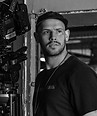 Cinematographer standing beside large camera rig on a film set backdrop.