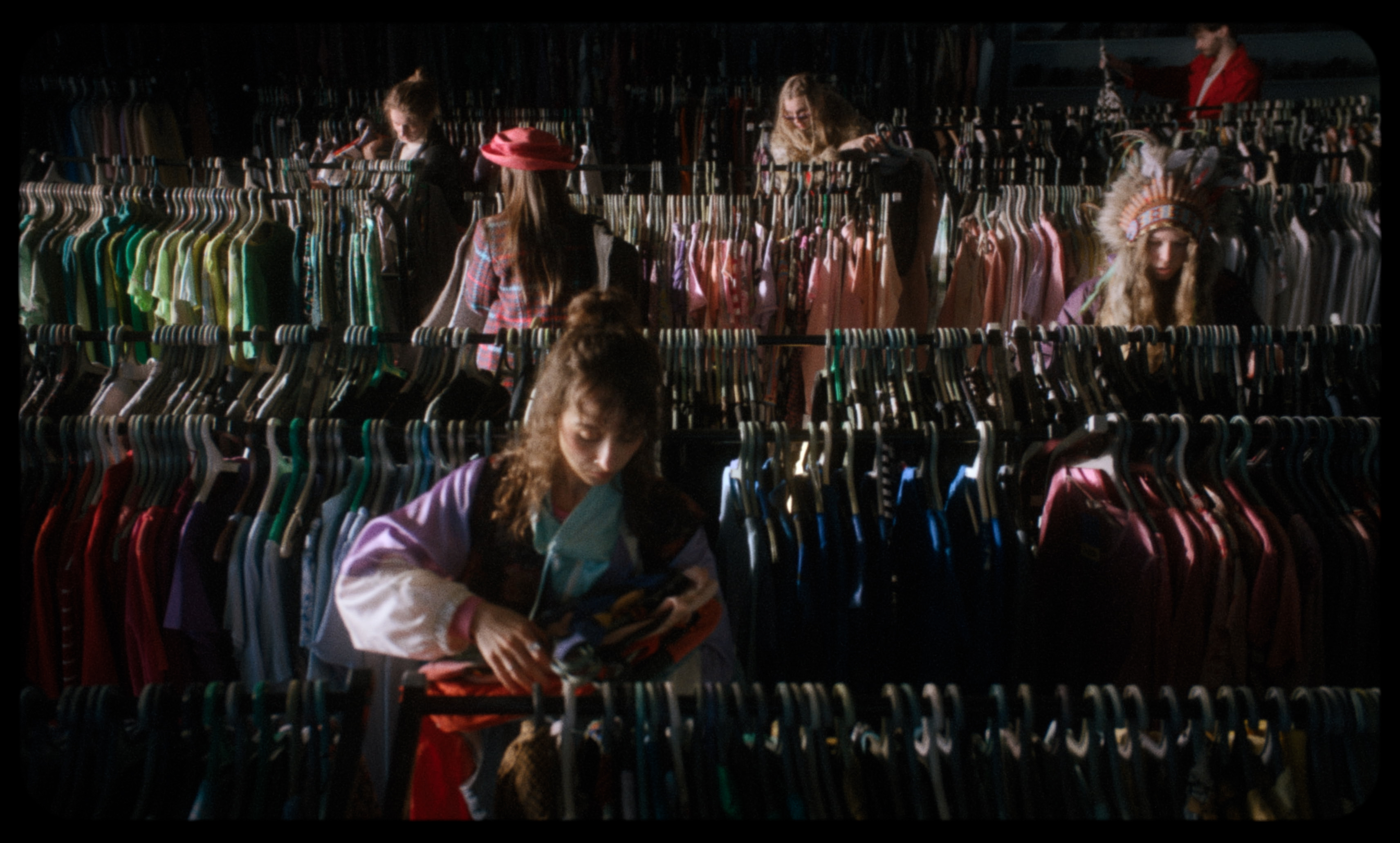 Young women browse clothing racks in a thrift store; one wears a feathered headdress — a still from